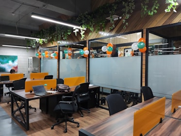 An open office space with wooden flooring and green plants hanging from the ceiling. There are multiple workstations with black chairs and orange partitions. Balloons in orange, white, and green are used for decoration, suggesting a festive or themed setup. Laptops and office supplies can be seen on the desks, and there is a painting on the wall to the left.