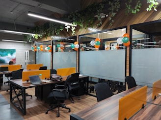 An open office space with wooden flooring and green plants hanging from the ceiling. There are multiple workstations with black chairs and orange partitions. Balloons in orange, white, and green are used for decoration, suggesting a festive or themed setup. Laptops and office supplies can be seen on the desks, and there is a painting on the wall to the left.