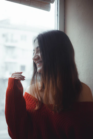 A serene shot of a woman in soft beige lounge wear, smiling gently by a sunlit window.