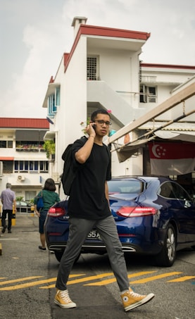 A person wearing glasses and casual clothing is walking in an urban area while talking on a smartphone. They are carrying a black backpack and wearing yellow high-top sneakers. In the background, there is a residential building with red and white colors and a blue car parked nearby. Several other people are visible, engaged in their own activities.