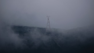 A tall, thin structure resembling a communication tower stands on a foggy, shadowy hill. The tower is partially obscured by dense fog that envelops the landscape, creating a mysterious and eerie atmosphere.