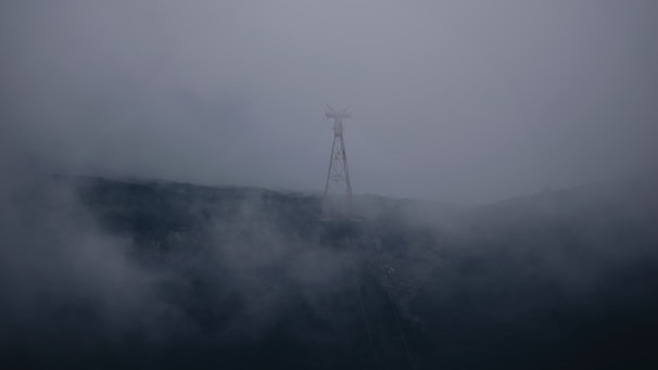 A tall, thin structure resembling a communication tower stands on a foggy, shadowy hill. The tower is partially obscured by dense fog that envelops the landscape, creating a mysterious and eerie atmosphere.