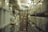 A long aisle in a warehouse or storage facility with shelves on both sides filled with boxes and containers. The shelves appear to be made of metal and are stacked high with various sized cardboard boxes and plastic bins. The lighting is dim, contributing to a slightly murky atmosphere.