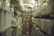 A long aisle in a warehouse or storage facility with shelves on both sides filled with boxes and containers. The shelves appear to be made of metal and are stacked high with various sized cardboard boxes and plastic bins. The lighting is dim, contributing to a slightly murky atmosphere.