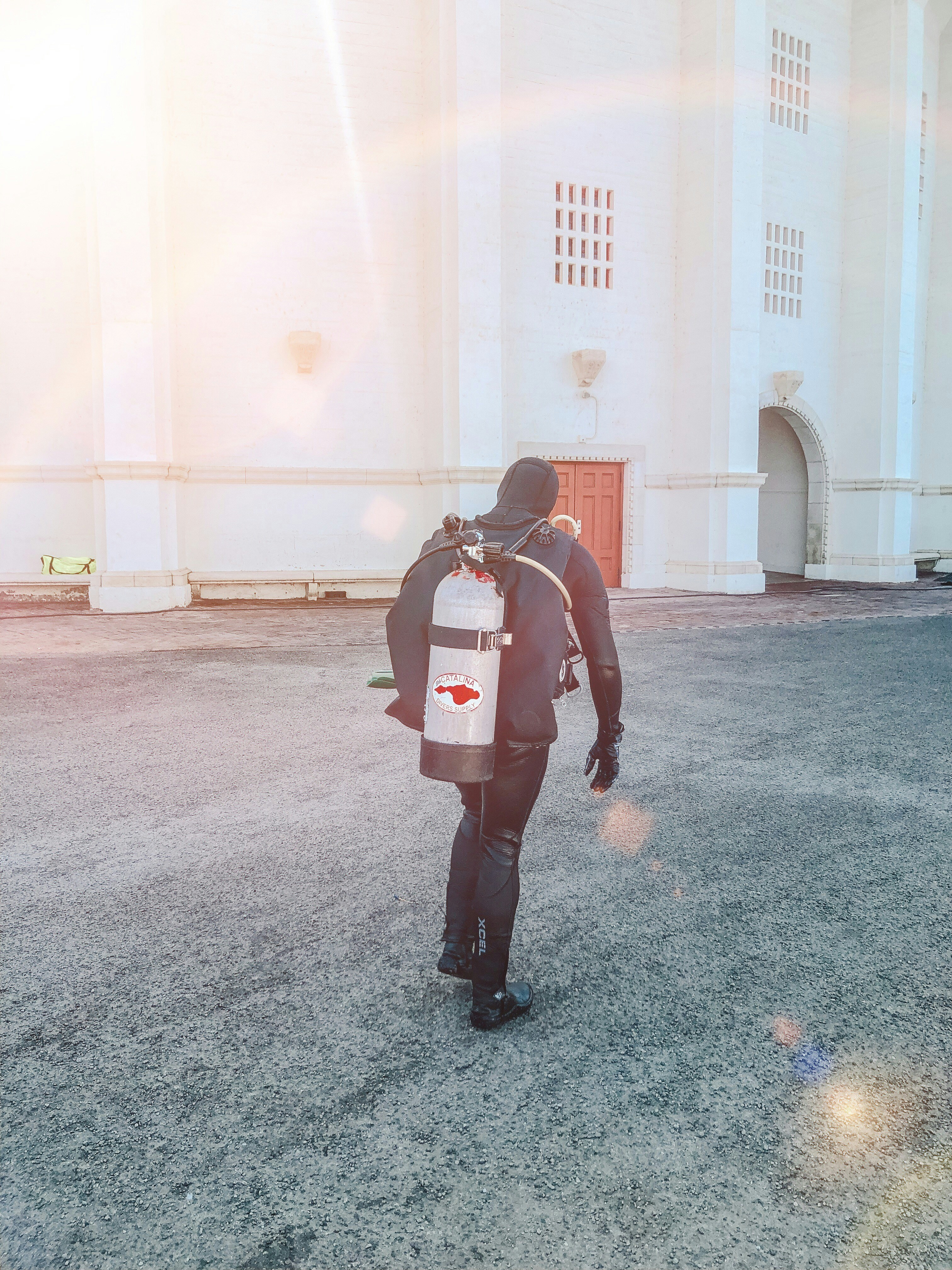 A person wearing a full diving suit, including a mask and fins, is carrying a large oxygen tank with a red fish logo on their back. The scene is set against a bright white building with red doors and an arched entrance, bathed in warm sunlight.