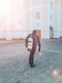 A person wearing a full diving suit, including a mask and fins, is carrying a large oxygen tank with a red fish logo on their back. The scene is set against a bright white building with red doors and an arched entrance, bathed in warm sunlight.