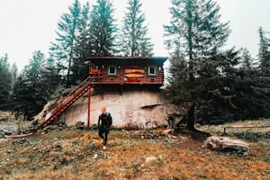 A wooden cabin perched atop a large rock, surrounded by tall evergreen trees. A person wearing outdoor gear and an orange hat stands in the foreground on grassy terrain. The cabin has a small balcony accessible by a staircase.
