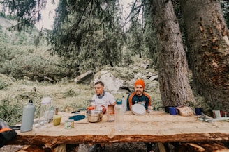Two people are sitting at a rustic wooden table in a forest setting, surrounded by trees and greenery. The table is covered with various camping items like bottles, containers, and cooking utensils. One person is wearing a bright orange beanie, while the other has a blue scarf around their neck. The scene conveys a casual outdoor camping experience.