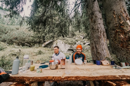 Two people are sitting at a rustic wooden table in a forest setting, surrounded by trees and greenery. The table is covered with various camping items like bottles, containers, and cooking utensils. One person is wearing a bright orange beanie, while the other has a blue scarf around their neck. The scene conveys a casual outdoor camping experience.