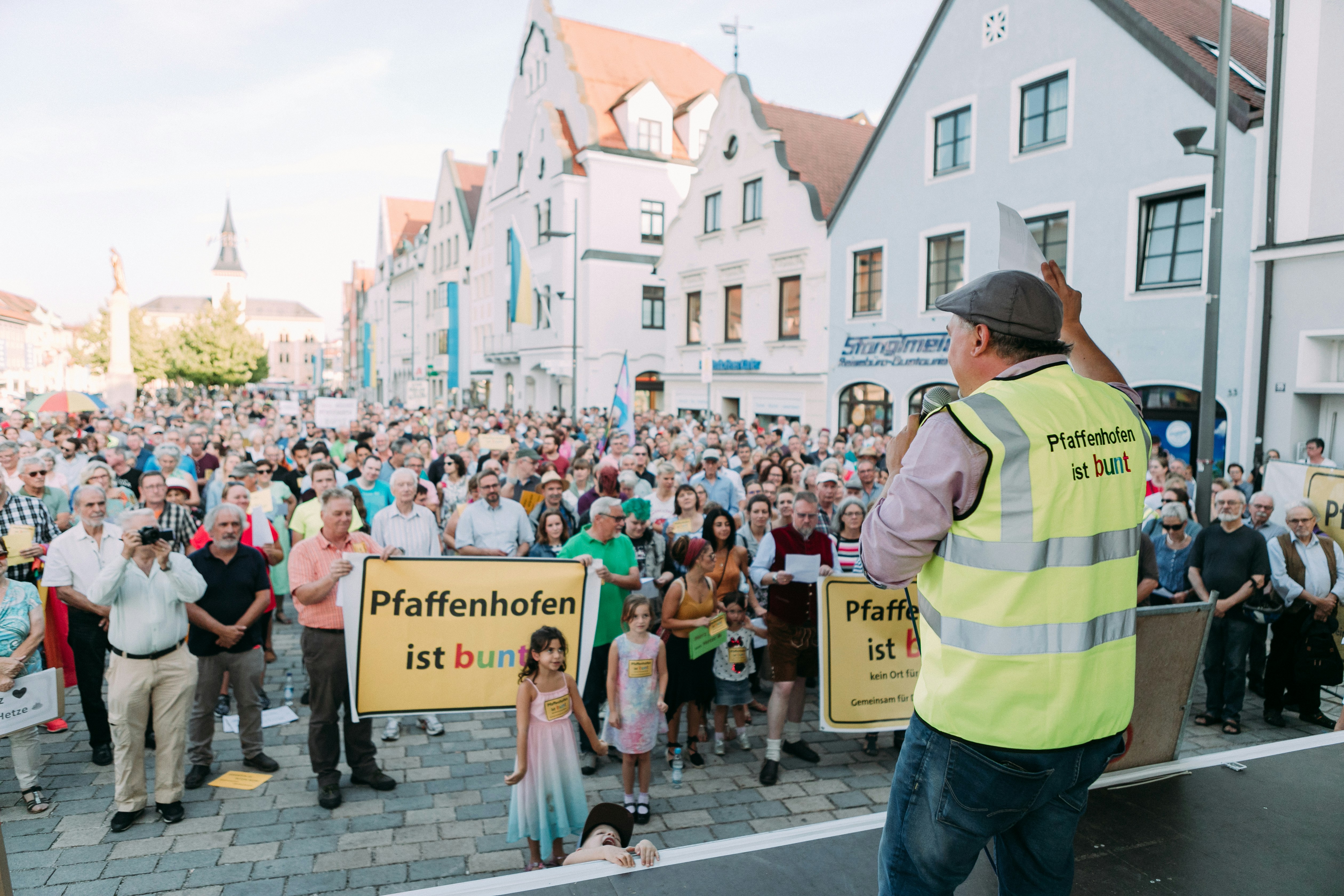 A speaker in a yellow vest addresses a crowd holding banners.