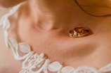 Close-up of two gold wedding rings resting on a person's collarbone, accompanied by intricate lace fabric with floral patterns, likely part of a bridal dress.