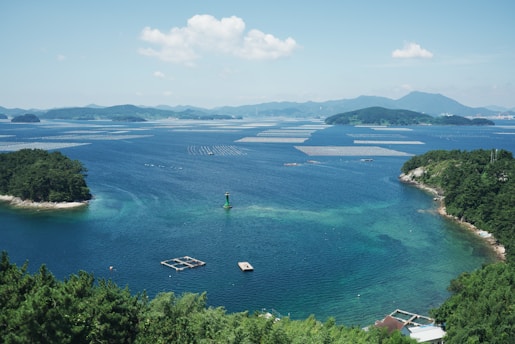 A serene coastal scene with clear blue waters and scattered islands covered in lush greenery. Floating platforms or fish farms are visible in the water, and a green marker buoy is positioned near the foreground. The distant background features a range of softly undulating mountains under a bright, partly cloudy sky.