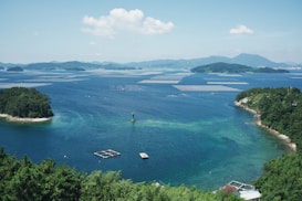 A serene coastal scene with clear blue waters and scattered islands covered in lush greenery. Floating platforms or fish farms are visible in the water, and a green marker buoy is positioned near the foreground. The distant background features a range of softly undulating mountains under a bright, partly cloudy sky.