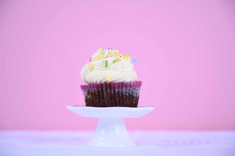 A beautifully decorated cupcake with creamy frosting and colorful sprinkles atop a white pedestal against a soft pink background.