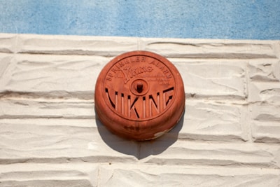 A red circular fire alarm, marked with 'Viking', is mounted on a textured, light beige brick wall. Above the alarm, the words 'Sprinkler Alarm' are visible, and the blue sky is in the background.