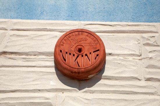 A red circular fire alarm, marked with 'Viking', is mounted on a textured, light beige brick wall. Above the alarm, the words 'Sprinkler Alarm' are visible, and the blue sky is in the background.