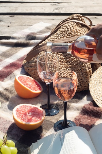 A picnic setup on a plaid blanket featuring two glasses being filled with rose wine, a woven straw bag, halved grapefruits, and a book. Sunlight casts warm shadows, creating a relaxed atmosphere.