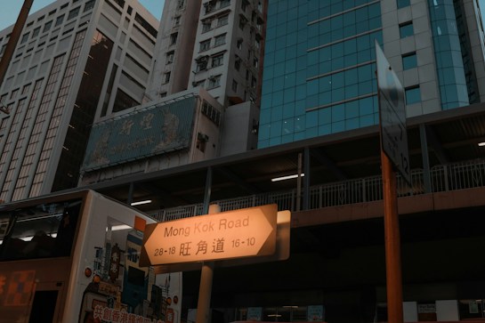 A bustling urban street scene with tall modern buildings, a road sign indicating Mong Kok Road, and part of a double-decker bus. The buildings have a mixture of glass facades and concrete, creating a diverse architectural skyline. The lighting gives an evening ambiance.