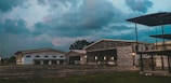 An open-air view of an airport hangar area with a few aircraft parked outside. The sky is covered with dark clouds creating a moody atmosphere, and the buildings have large doors suggesting a hub for storing airplanes or conducting related business.