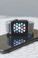 A smartwatch with a gray woven band is positioned on top of a smartphone with a black screen, which reflects the watch's face displaying multiple colorful app icons.