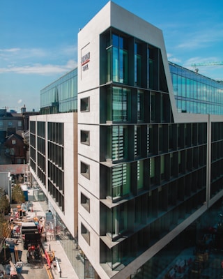 A modern multi-story building with large glass windows and a distinctive sloping architectural design. The structure is surrounded by some smaller traditional buildings in the background. Visible on the street below are construction materials and vehicles, including a truck and some scaffolding.