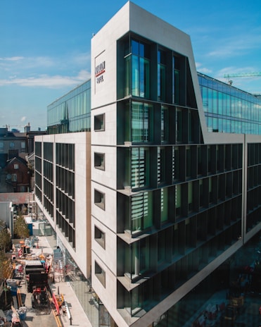 A modern multi-story building with large glass windows and a distinctive sloping architectural design. The structure is surrounded by some smaller traditional buildings in the background. Visible on the street below are construction materials and vehicles, including a truck and some scaffolding.