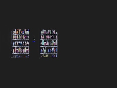 Two vending machines filled with various snacks and beverages are illuminated against a dark background. The machines contain items such as chips, candy bars, and bottled drinks organized neatly in rows.