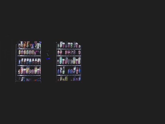 Two vending machines filled with various snacks and beverages are illuminated against a dark background. The machines contain items such as chips, candy bars, and bottled drinks organized neatly in rows.