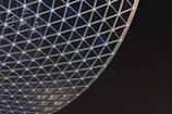 A large geodesic dome structure with a pattern of triangular glass panels and illuminated intersecting points creating a lattice-like design. The background is a dark sky, which highlights the bright lights at the nodes of the structure.