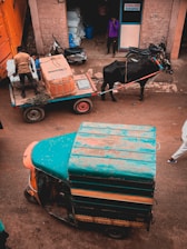 A street scene featuring a cow-drawn cart carrying large parcels, supervised by a man. This is positioned near a vibrant auto rickshaw with worn paintwork. The background includes a partially open storefront and several motorbikes parked nearby. Another man is standing at the entrance of the store, talking on a phone.