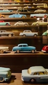 A collection of miniature vintage cars displayed on dark wooden shelves. The cars vary in color and design, including shades of blue, green, red, and black. Each vehicle is intricately detailed, capturing a nostalgic charm.