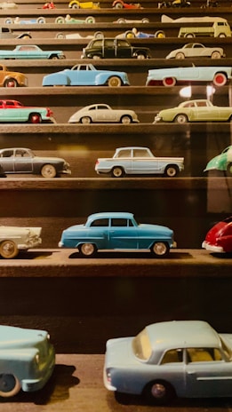 A collection of miniature vintage cars displayed on dark wooden shelves. The cars vary in color and design, including shades of blue, green, red, and black. Each vehicle is intricately detailed, capturing a nostalgic charm.