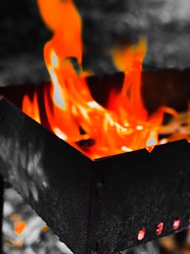 Bright orange and yellow flames rise from a black metal container, creating a contrast with the muted background. The metal surface is visibly textured and dark, providing a stark backdrop to the vivid colors of the fire.