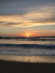 The image displays a serene beach at sunset with gentle waves crashing onto the shore. The sky is a blend of warm hues, transitioning from vibrant orange and yellow near the horizon to cooler blues and grays as it rises.