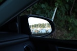 A reflection of several boats is visible in a car’s side mirror. The boats are docked on a body of water, surrounded by trees and a clear sky. The area around the mirror is comprised of car interior and green foliage.