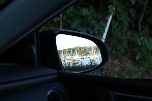 A reflection of several boats is visible in a car’s side mirror. The boats are docked on a body of water, surrounded by trees and a clear sky. The area around the mirror is comprised of car interior and green foliage.