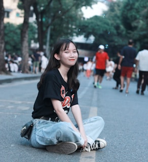 A young person sits cross-legged on a street, smiling slightly. They are wearing a black T-shirt with graphic designs and light blue jeans. In the background, people walk along the street lined with trees, creating a casual and relaxed urban atmosphere.