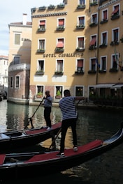 Two gondoliers navigate their gondolas along a canal in front of a yellow building with red awnings. The building is adorned with flower boxes under the windows and has a sign that reads 'Hotel Cavalletto.' The water reflects the gondolas and the architecture, creating a picturesque scene.