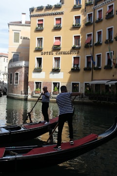 Two gondoliers navigate their gondolas along a canal in front of a yellow building with red awnings. The building is adorned with flower boxes under the windows and has a sign that reads 'Hotel Cavalletto.' The water reflects the gondolas and the architecture, creating a picturesque scene.