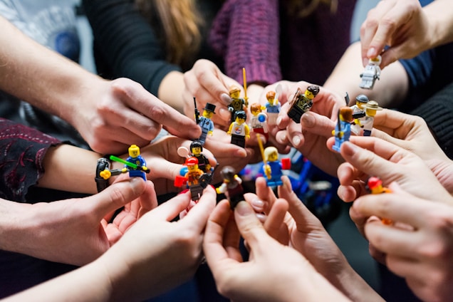 Multiple hands, mostly caucasian, are holding a variety of colorful LEGO figurines in a playful manner, each person appearing to display a different figurine.