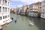 A scenic canal with traditional gondolas and a motorboat navigating through the water. Historic buildings with classical architecture line both sides of the canal, featuring arched windows and pastel-colored facades. The sky is clear with a few clouds, indicating a bright, sunny day.