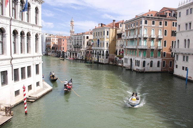 A scenic canal with traditional gondolas and a motorboat navigating through the water. Historic buildings with classical architecture line both sides of the canal, featuring arched windows and pastel-colored facades. The sky is clear with a few clouds, indicating a bright, sunny day.