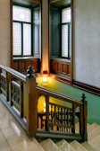 A beautifully designed staircase with ornate wooden railing and intricate stained glass windows. The wooden craftsmanship is detailed and complements the warm lighting fixtures on the walls, creating a vintage or historical atmosphere.
