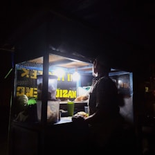 A person is preparing food inside a dimly-lit street food stall at night. The stall is illuminated by a single bright light bulb, revealing containers and food supplies. The signs display menu items and prices.