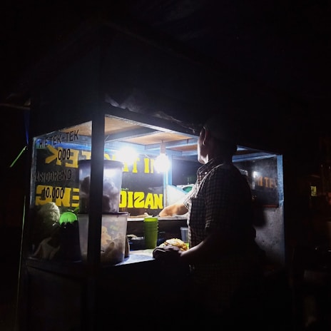 A person is preparing food inside a dimly-lit street food stall at night. The stall is illuminated by a single bright light bulb, revealing containers and food supplies. The signs display menu items and prices.