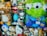 A display of various plush toys in a store window, including characters from popular animations. The toys are a mix of animals and cartoon figures, some of which are wrapped in plastic. Among them are a large green alien with big eyes, a brown bear, and several other colorful stuffed animals.