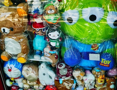 A display of various plush toys in a store window, including characters from popular animations. The toys are a mix of animals and cartoon figures, some of which are wrapped in plastic. Among them are a large green alien with big eyes, a brown bear, and several other colorful stuffed animals.