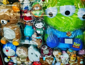 A display of various plush toys in a store window, including characters from popular animations. The toys are a mix of animals and cartoon figures, some of which are wrapped in plastic. Among them are a large green alien with big eyes, a brown bear, and several other colorful stuffed animals.