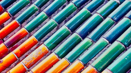 Rows of colorful ceramic tiles installed diagonally. The tiles display a gradient of colors ranging from vibrant orange to deep green. Each tile has a glossy finish, reflecting light in a playful manner.