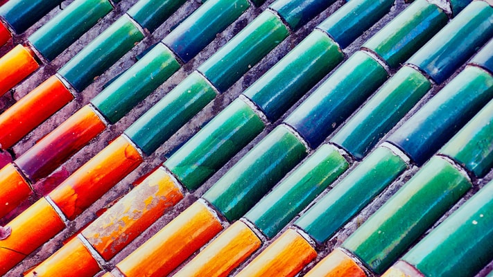 Rows of colorful ceramic tiles installed diagonally. The tiles display a gradient of colors ranging from vibrant orange to deep green. Each tile has a glossy finish, reflecting light in a playful manner.